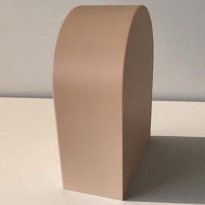 May include: A solid, light beige, arch-shaped object with a smooth, matte finish. The piece features a flat base, vertical sides, and a rounded top, standing upright as a singular, three-dimensional form with clean, defined edges and surfaces.