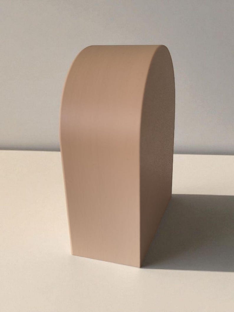 May include: A solid, light beige, arch-shaped object with a smooth, matte finish. The piece features a flat base, vertical sides, and a rounded top, standing upright as a singular, three-dimensional form with clean, defined edges and surfaces.