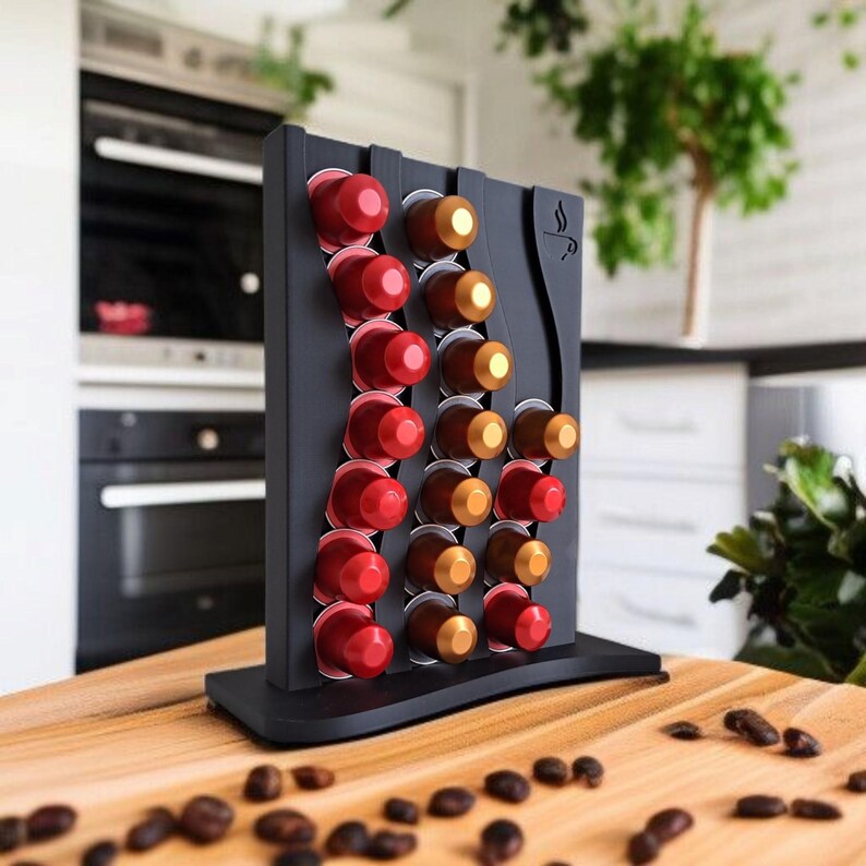 May include: A black vertical coffee pod holder with a wavy design, featuring a small coffee cup icon. The stand is filled with rows of red and gold coffee capsules. The holder is positioned on a wooden surface scattered with dark roasted coffee beans.