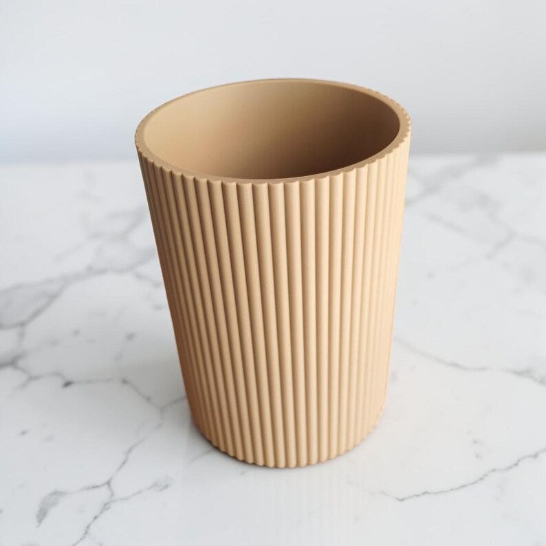 May include: A cylindrical wastebasket or storage container in a solid light beige color. The exterior surface features a consistent vertical ribbed or fluted texture that runs from the top rim to the base, creating a uniform pattern around the object.