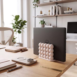 May include: A dark gray Dell laptop held upright by a light pink, bubble-textured vertical stand. The stand features a grid of rounded, sphere-like shapes on a flat base, providing a stable support for the device on a wooden desk surface.