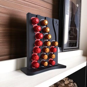 May include: A matte black vertical coffee pod holder with a wavy design, featuring three columns of slots filled with red and gold coffee capsules. The top right corner displays a small engraved icon of a coffee cup with steam rising from it.