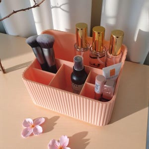 May include: A peach-coloured plastic makeup organiser with vertical ribbed detailing and multiple compartments. It contains two makeup brushes, three clear bottles with gold caps, a black spray bottle, and two small tubes of skincare products.
