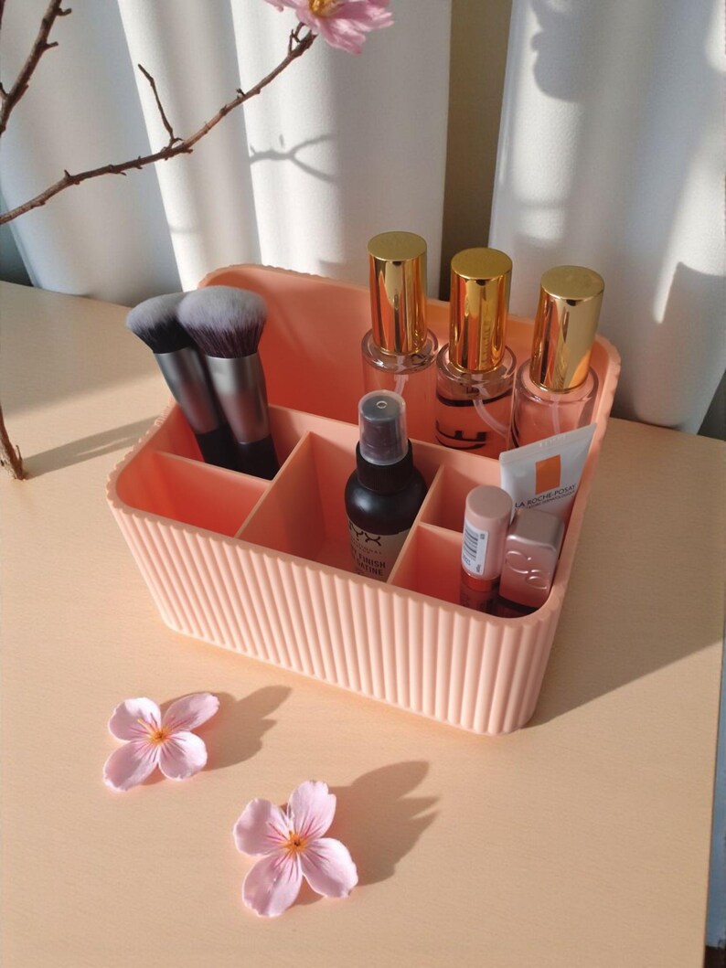 May include: A peach-coloured plastic makeup organiser with vertical ribbed detailing and multiple compartments. It contains two makeup brushes, three clear bottles with gold caps, a black spray bottle, and two small tubes of skincare products.