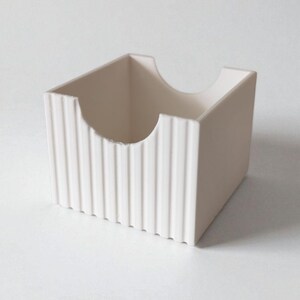 May include: A square, off-white storage container featuring a ribbed, vertical texture on its exterior walls. The front panel includes a semi-circular cutout at the top edge, providing an open access point to the interior of the box.