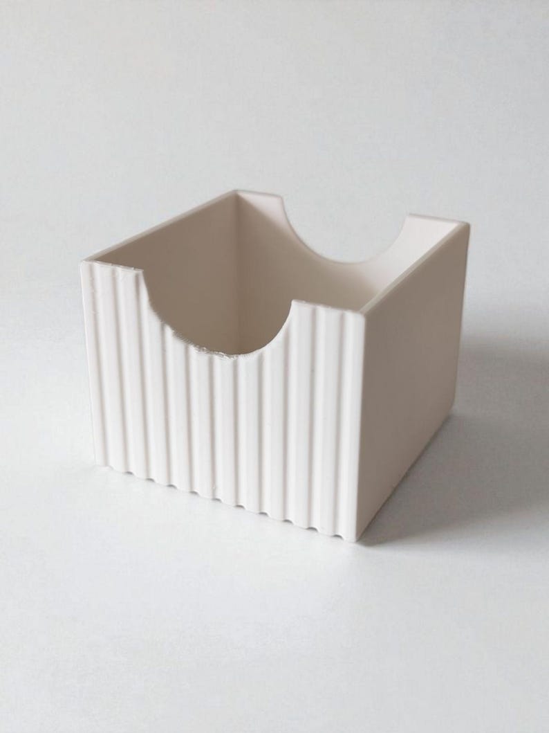 May include: A square, off-white storage container featuring a ribbed, vertical texture on its exterior walls. The front panel includes a semi-circular cutout at the top edge, providing an open access point to the interior of the box.