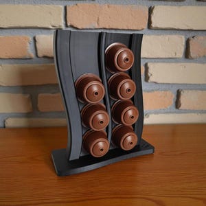 Puede incluir: Un soporte vertical negro para c&aacute;psulas de caf&eacute; con dise&ntilde;o ondulado, que cuenta con dos columnas para albergar un total de ocho c&aacute;psulas de caf&eacute; marrones. El soporte tiene una base plana y un acabado mate liso para organizar c&aacute;psulas.