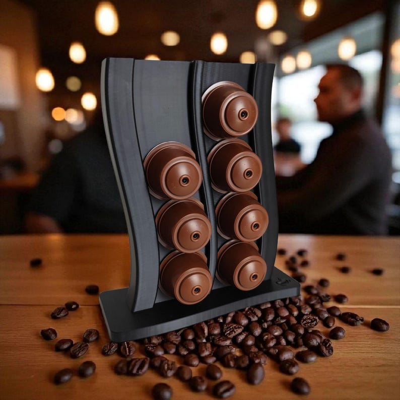 Puede incluir: Un soporte vertical negro para c&aacute;psulas de caf&eacute; con dise&ntilde;o ondulado, que cuenta con dos columnas para un total de ocho c&aacute;psulas de caf&eacute; marrones. El soporte est&aacute; colocado en posici&oacute;n vertical sobre una superficie plana con granos de caf&eacute;.