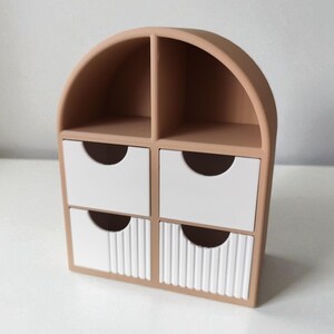 May include: A tan desktop organizer with an arched top, featuring two open upper compartments and four lower drawers. The two bottom drawers have a vertical ribbed texture on their white faces, while the two top drawers are smooth white.