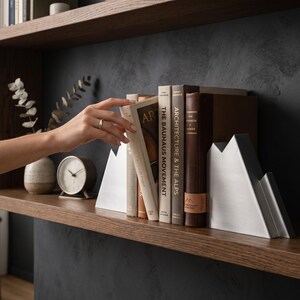 Peut inclure: Une paire de serre-livres en m&eacute;tal argent&eacute; en forme de sommets montagneux, plac&eacute;s sur une &eacute;tag&egrave;re en bois pour soutenir une collection de livres reli&eacute;s, dont les titres The Bauhaus Movement et Architecture and the Alps sont visibles.