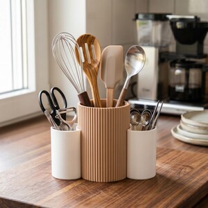 May include: A kitchen utensil holder featuring a central ribbed tan cylinder flanked by two smaller smooth white cylindrical containers. The holder contains a metal whisk, a wooden slotted spoon, a silicone spatula, a metal ladle, scissors, and a peeler.
