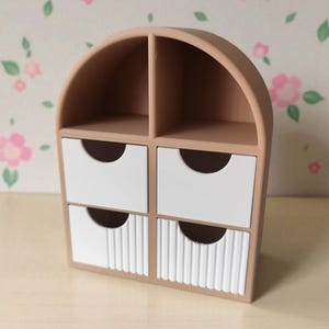 May include: A beige desktop organizer with an arched top, featuring two open upper compartments and four lower drawers. The two top drawers have smooth white fronts, while the two bottom drawers feature a vertical ribbed texture on the white fronts.