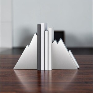 Peut inclure: Une paire de serre-livres en m&eacute;tal argent&eacute; en forme de sommets de montagne, plac&eacute;s de chaque c&ocirc;t&eacute; de trois livres verticaux. Les serre-livres pr&eacute;sentent une finition m&eacute;tallique bross&eacute;e avec des silhouettes angulaires triangulaires.