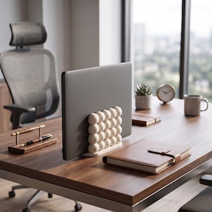May include: A dark gray laptop held upright by a white stand featuring a grid of sixteen spherical protrusions. The stand is positioned on a wooden desk surface alongside a brown leather-bound notebook, a pen on a stand, a small plant, and a clock.