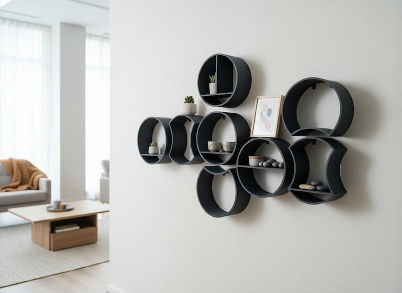 May include: A collection of matte black circular wall shelves arranged in a cluster. The shelves feature various internal dividers and horizontal surfaces holding small potted succulents, ceramic bowls, smooth stones, and a framed picture.