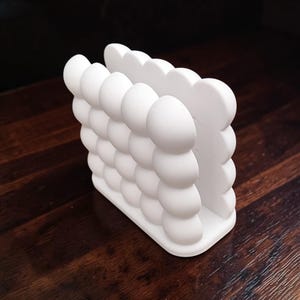 May include: A white napkin holder featuring a repeating bubble or sphere pattern on both sides. The object has a rectangular base and a vertical slot for holding napkins, constructed with a matte finish and a uniform, rounded, three-dimensional texture.