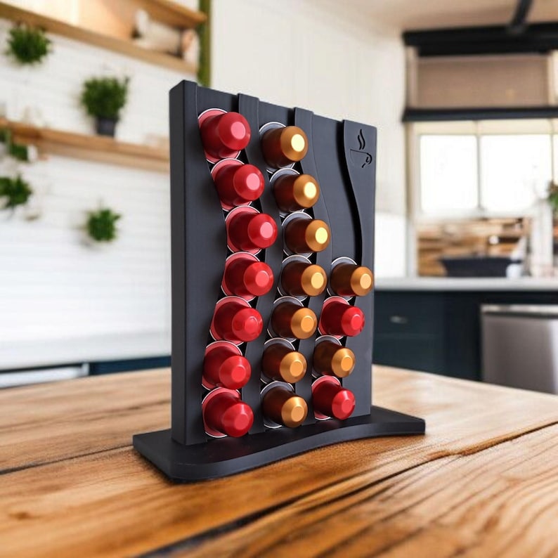 May include: A black vertical coffee pod holder with a wavy design, featuring three columns of slots filled with red and gold coffee capsules. The stand has a flat base and a small coffee cup icon engraved on the upper right side of the frame.