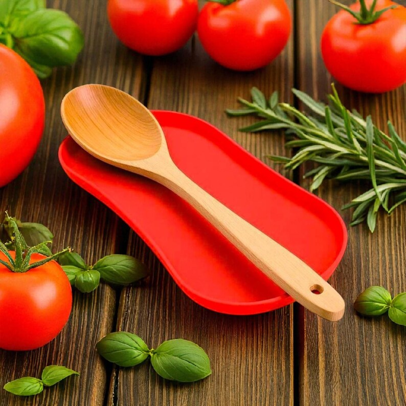 May include: A light-coloured wooden spoon resting on a bright red oval silicone spoon rest. The spoon has a smooth, rounded bowl and a handle with a small circular hole at the end. The spoon rest is solid red with a smooth, matte-like surface.