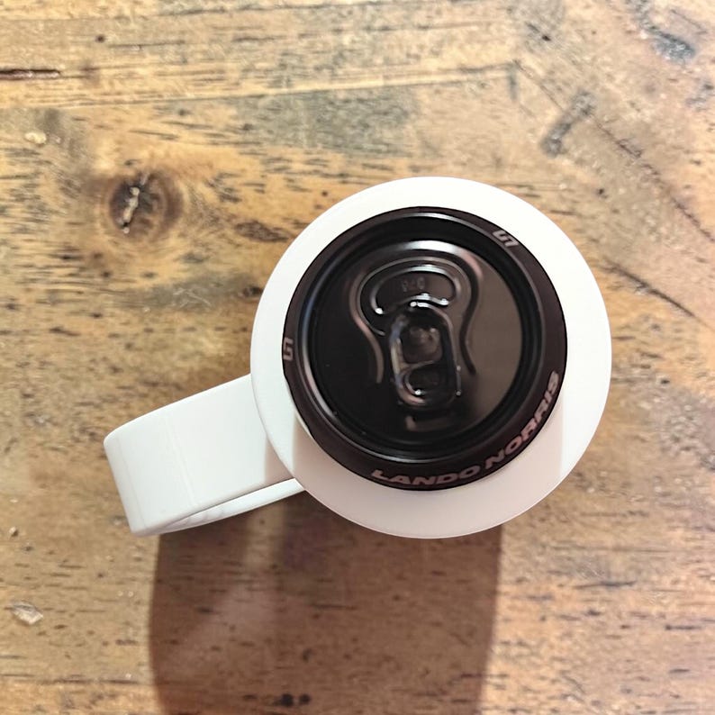 May include: A white beverage holder with a handle, featuring a black top lid with a pull tab. The black lid is printed with the text Lando Norris and the number 4. The design is viewed from directly above, showing the circular shape and handle.