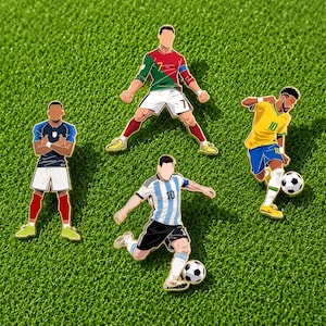 Puede incluir: Cuatro pines de esmalte que representan a jugadores de fútbol con diferentes uniformes. Un jugador viste una camiseta roja y verde con pantalón blanco, otro una camiseta azul con pantalón blanco, una camiseta amarilla con pantalón azul y una a rayas.