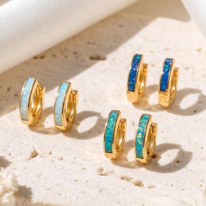 May include: Three pairs of small gold hoop earrings featuring inlaid stone accents. One pair has light blue stone, one pair has dark blue stone, and one pair has teal stone. The earrings are circular with a polished gold finish and a hinged closure.