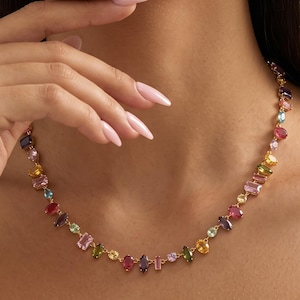 May include: A gold chain necklace featuring a series of multi-coloured gemstones in various shapes, including oval, rectangular, and marquise cuts. The stones display a vibrant palette of pink, yellow, green, blue, and purple hues set in gold.