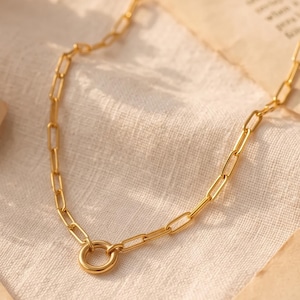 Op de afbeelding: Een goudkleurige ketting met langwerpige rechthoekige schakels en een enkele ronde ringverbinding in het midden. De metalen ketting heeft een gepolijste afwerking en is in een boogvorm gerangschikt op een lichtgekleurde stoffen ondergrond.