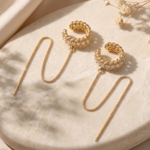 May include: A pair of gold-toned ear cuffs featuring a textured chain-link design embedded with small clear stones. Each cuff is attached to a delicate, thin gold-toned chain that hangs down, ending in a straight, slender metal post.
