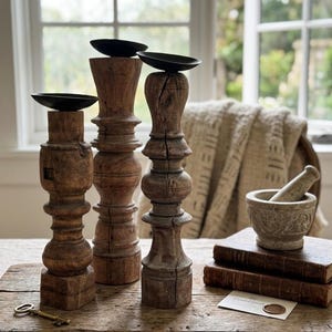 May include: Three tall, turned wooden candle holders with dark metal plates at the top. The wood has a weathered, brown finish with visible grain and cracks. Each holder features a distinct, carved silhouette with rounded and square base sections.