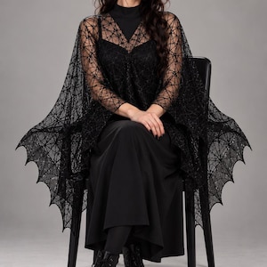 May include: A black sheer cape featuring a geometric spiderweb pattern and scalloped bat-wing edges. The cape is worn over a solid black sleeveless top and a long black skirt, paired with black lace-up combat boots with thick lug soles.