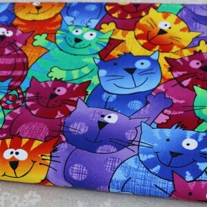 Puede incluir: Una tela con un patrón repetitivo de gatos de dibujos animados en colores vibrantes que incluyen azul, morado, verde, naranja, rojo y amarillo. Cada gato tiene una expresión distinta, ojos grandes y una apariencia texturizada con detalles.