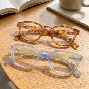May include: Two pairs of spectacles with rounded frames. The top pair features a multi-coloured tortoiseshell pattern in shades of brown, orange, and green. The bottom pair has a translucent, light blue-tinted frame with a smooth finish.