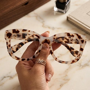 May include: Spectacles with a thick, translucent frame featuring a brown and cream tortoiseshell pattern. The glasses have a rounded, oversized shape with clear lenses and a distinct bridge connecting the two rims of the frame.