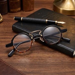 May include: Round eyeglasses featuring a tortoiseshell and black frame with a gold-toned metal bridge detail. The glasses have clear lenses and are positioned in front of a black fountain pen resting on a black leather-bound notebook.
