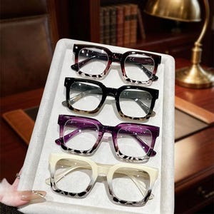 May include: Four pairs of thick-rimmed square glasses arranged vertically. The frames feature a gradient design, transitioning from solid colours at the top to a spotted animal print pattern at the bottom in shades of burgundy, black, purple, and cream.