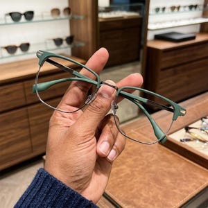 May include: Eyeglasses featuring a matte sage green browline frame with thin silver metal rims. The design includes a bridge and temples in a matching sage green tone, with small silver accents at the outer corners of the frame front.
