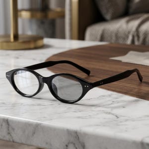 May include: A pair of black-framed spectacles with oval-shaped lenses. The frames feature a glossy finish and two small silver-toned rivets on each side near the hinges. The temples are straight and extend outward from the front of the frames.