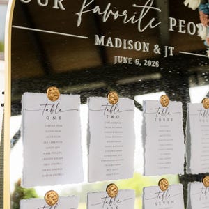 Peut inclure: Un plan de table en acrylique noir en forme d'arche avec un cadre doré, portant le texte blanc "OUR favorite PEOPLE", "MADISON & JT" et "JUNE 6, 2026". Des cartes en papier blanc à bords déchirés sont fixées par des épingles dorées.