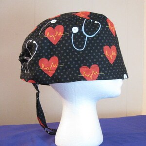 May include: Black scrub hat with a white stethoscope and red hearts with yellow EKG lines on a black background with white polka dots.