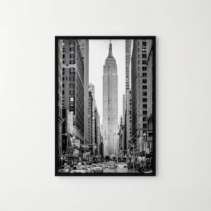 Könnte beinhalten: Ein gerahmter Schwarz-Weiß-Wandkunstdruck, der eine vertikale Perspektive einer Straßenschlucht in New York City zeigt. Das Empire State Building steht markant in der Mitte, eingerahmt von hohen Wolkenkratzern auf beiden Seiten, mit Verkehr darunter.