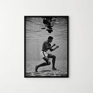 May include: A black and white framed photograph depicting a boxer underwater in a swimming pool. The boxer is in a crouched stance, wearing white trunks with dark side stripes, with their reflection visible on the water surface above.