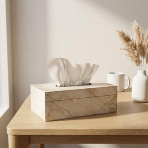 Puede incluir: Una caja para pañuelos rectangular con un patrón de imitación mármol en tonos beige claro y bronceado con acabado liso. Un pañuelo blanco sale por la abertura superior, extendiéndose hacia arriba en una forma suave y plegada sobre la caja.