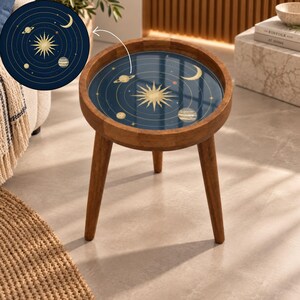 May include: A round wooden side table with three tapered legs and a circular tabletop featuring a dark blue inlay. The inlay displays a gold celestial design with a central sun, various planets, stars, and a crescent moon in a solar system layout.