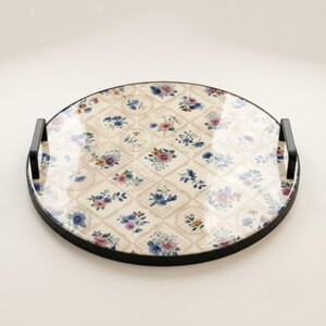 May include: A round serving tray with a black metal frame and two black handles. The surface features a cream-colored background with a repeating diamond pattern containing small, colorful floral illustrations in shades of blue, pink, and yellow.