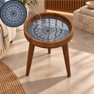 May include: A round wooden side table with three tapered legs and a circular tabletop featuring a glossy, intricate blue and white mandala pattern. The design displays a detailed, symmetrical floral motif centered on the flat surface.