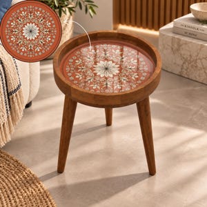 May include: A round wooden side table with three tapered legs and a circular tabletop featuring a glossy, terracotta-colored inlay. The inlay displays a symmetrical, white floral mandala pattern with intricate, repeating petal and vine motifs.