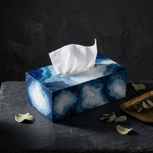 May include: A rectangular tissue box cover featuring a vibrant blue and white agate stone pattern with gold-toned veining details. A single white facial tissue is pulled through the top opening of the box, which displays a polished finish.
