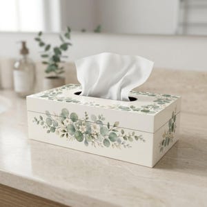Peut inclure: Une boîte à mouchoirs rectangulaire de couleur crème, ornée d'un motif botanique à l'aquarelle représentant des feuilles d'eucalyptus vertes et de petites fleurs blanches. Un mouchoir blanc dépasse de l'ouverture ovale sur le dessus.
