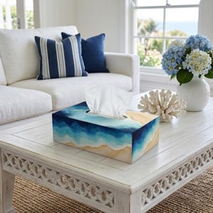 May include: A rectangular tissue box cover featuring a painted design of ocean waves crashing onto a sandy beach. The artwork displays shades of deep blue, turquoise, and white foam transitioning into a tan shoreline along the bottom edges.