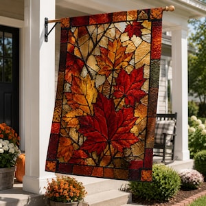May include: A rectangular garden flag featuring a stained glass mosaic pattern of autumn maple leaves in shades of deep red, burnt orange, and golden yellow, framed by a dark border with small leaf accents at the corners, hanging from a metal pole.